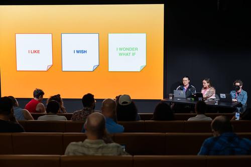 a presentation or workshop setting in an auditorium or conference room. Three speakers are seated at a table on stage at the right side, with audience members visible in the foreground seated in rows. The large projection screen displays three sticky notes with different colored borders (red, blue, and green) containing the phrases 'I LIKE,' 'I WISH,' and 'I WONDER WHAT IF' respectively.