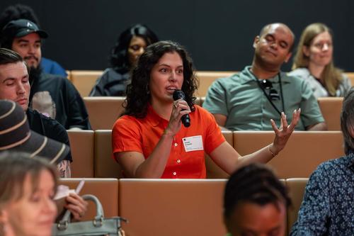 A woman with dark hair and a red shirt speaks into a microphone while sitting