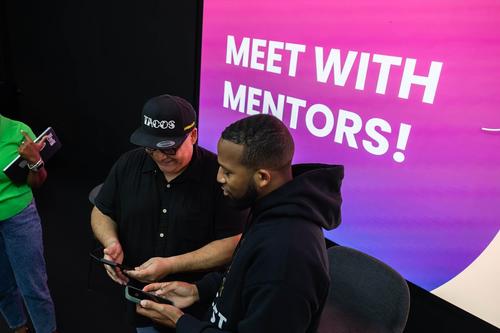 A Latino man with a hat and a Black man with short hair and a beard hold their phones together, standing in front of a projection of reading 'Meet with Mentors'