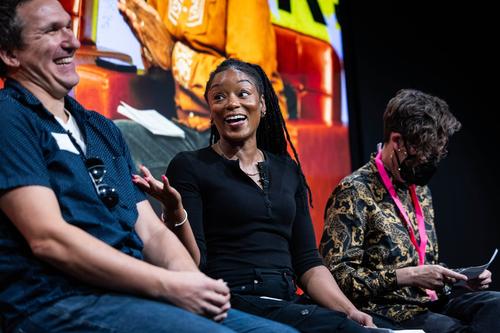 On a panel of speakers, a Black woman in a black top smiles and gestures towards a laughing white man in a blue shirt. A white woman in a mask sits next to her.