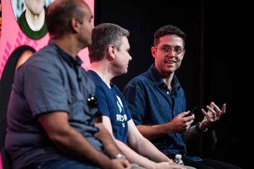 On a panel of speakers, a white man in glasses gestures as he speaks toward two other men in the foreground