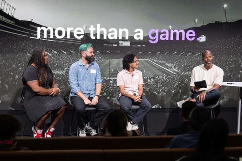 On a panel of spakers, a Black woman in a Black dress (Amira Rose Davis), a white man with green hair (Landon Cotham) and a Latino man in a white shirt (Juan Garcia) sit looking toward a Black man in a white shirt who is speaking. The screen projection behind them reads 'more than a game.'