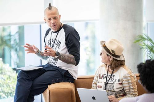 A white man with tatoos on his arm wearing a black and white shirt gestures as he sits on the arm of a couch. In the forground a white woman with a laptop and a hat looks toward him.