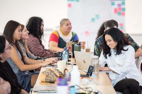 A group of seven people sit around a long table with laptops, water bottles and disposable cups.