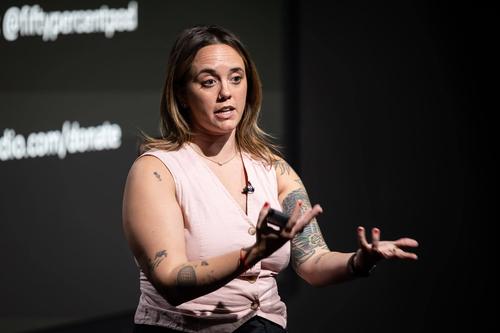 A white woman with tatoos on her arms and a pink vest gestures as she speaks in front of a screen.