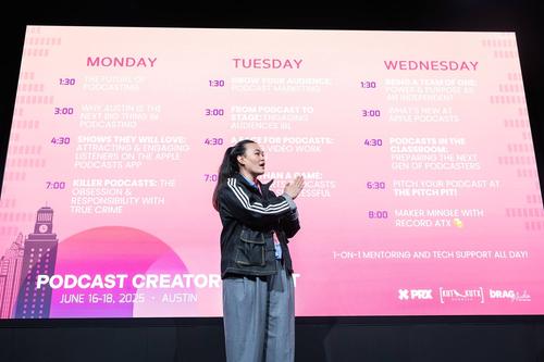 An East Asian woman with long hair in a ponytail and a black jacket stands in front of a screen projecting a timetable of events