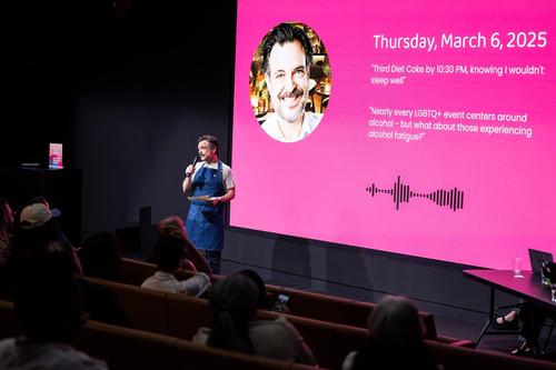 A white man with a mustache wearing an apron speaks toward a seated audience in front of a screen projection of his image.