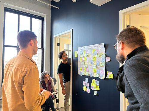A diverse group of people sits around whiteboard containing sticky notes as one member talks