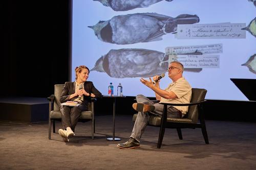 A woman and a man are seated in conversation on a stage in an auditorium.